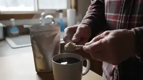 A man in his sixties tipping a small spoon of pale powder into a mug of black coffee at a kitchen counter